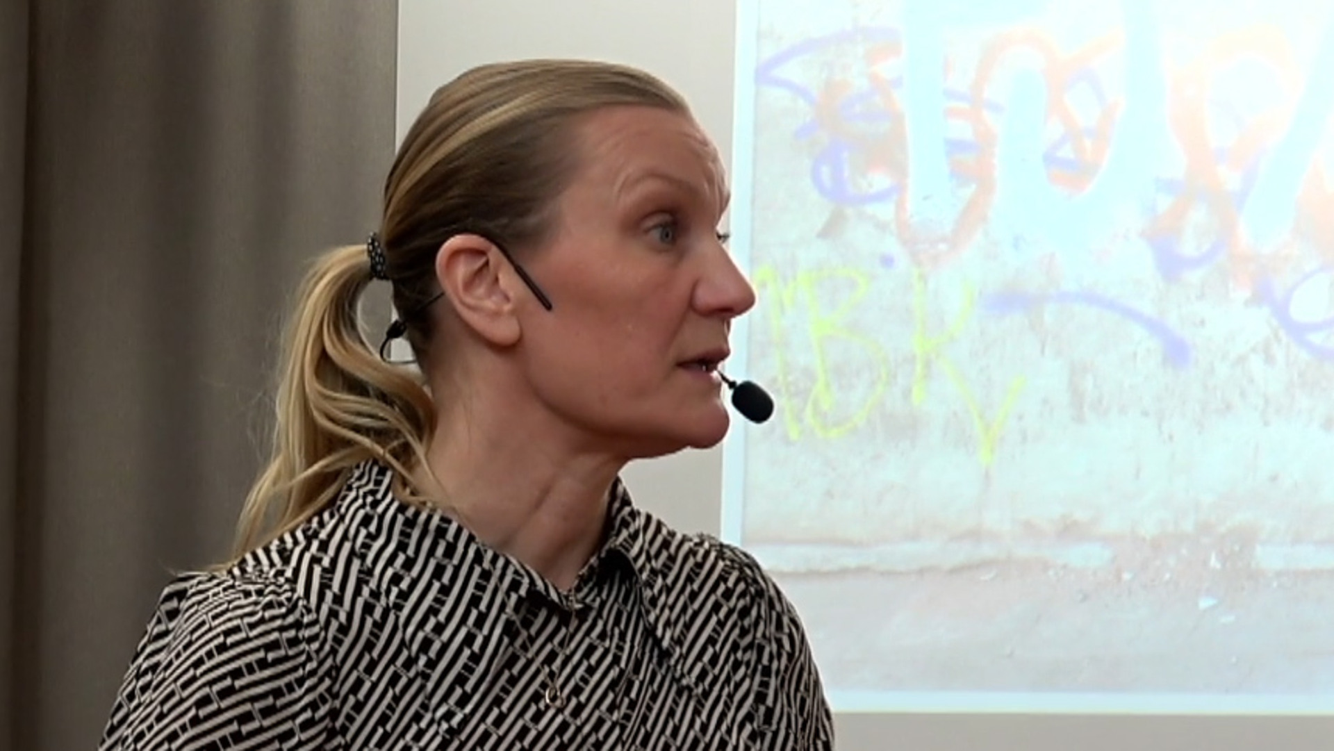 &Aring;sa Konradsson Geuken f&ouml;rel&auml;ser p&aring; infoteket. P&aring; projektorduken bakom visas en m&aring;lning av hennes bror Mats.