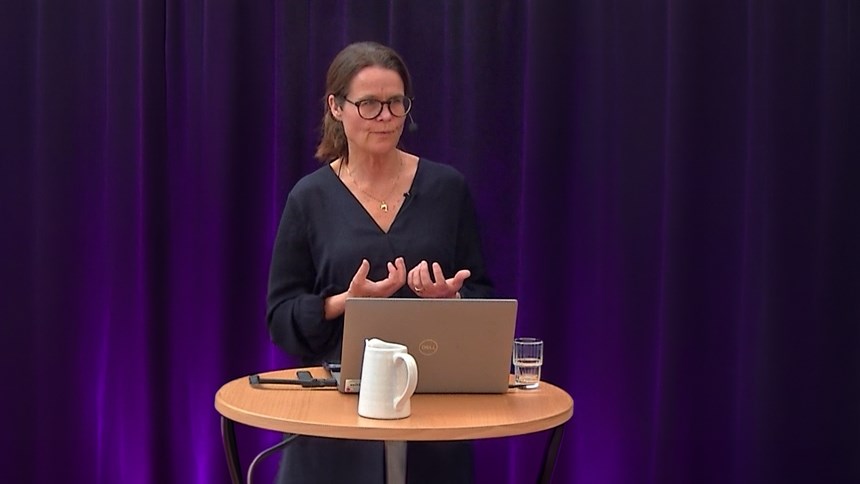 Anna Ehn gestikikulerar bakom ett st&aring;bord med f&ouml;rel&auml;sardator. I bakgrunden ett violett draperi.