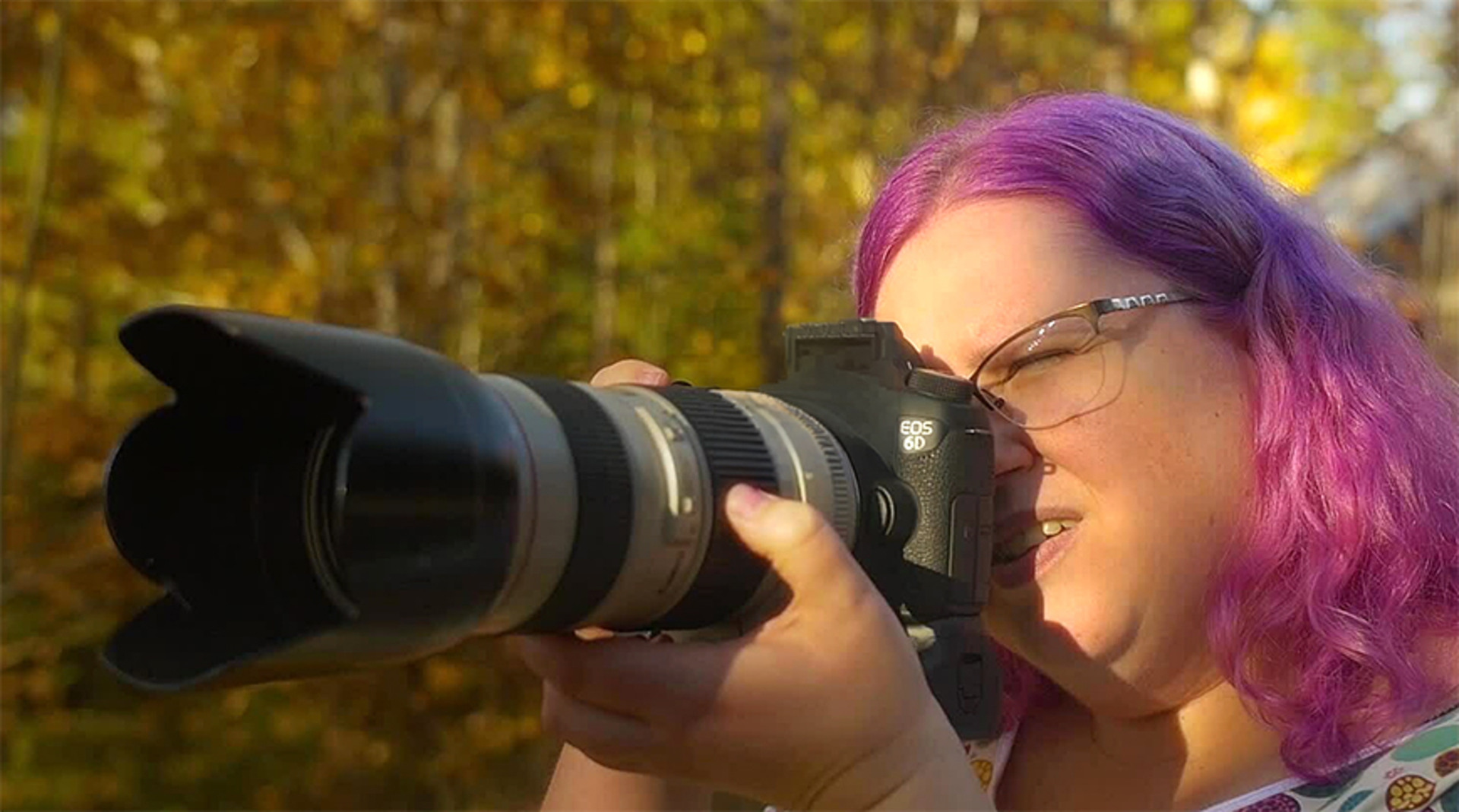 Person med l&aring;ng lilaf&auml;rgat h&aring;r tittar in i kamera som har ett l&aring;ngt objektiv.
