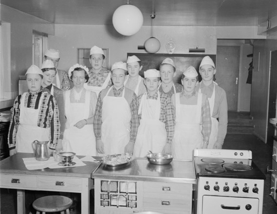 En gruppbild i köksmiljö med en lärare och ett tiotal yngre elever i förkläden.