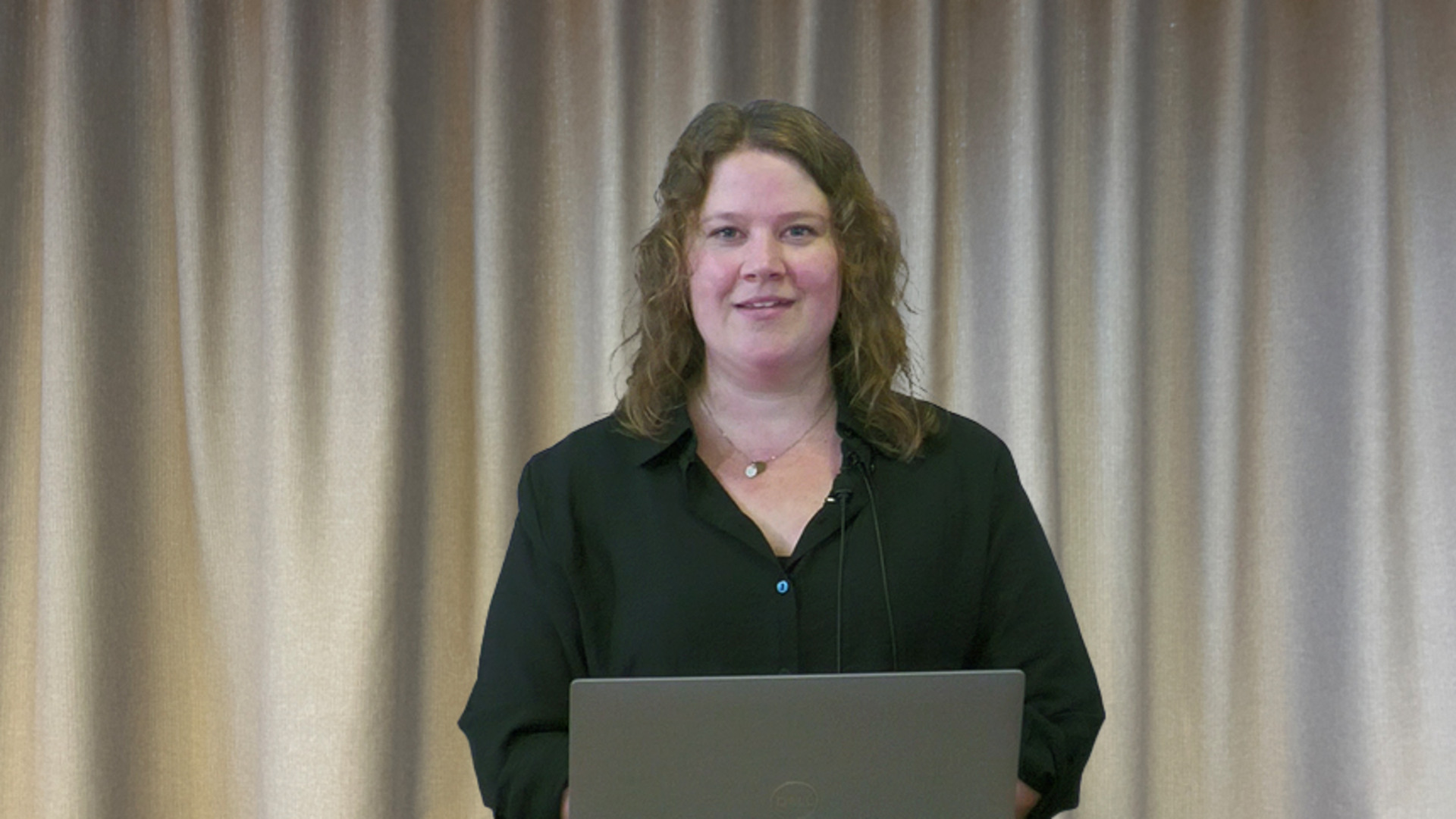 Jenny Wennerberg i svart blus, l&aring;ngt m&ouml;rkt h&aring;r vid laptop framf&ouml;r gr&aring;tt draperi.
