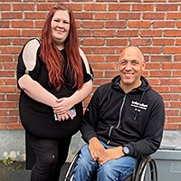 Tess st&aring;ende och Niklas sittande i rullstol, framf&ouml;r tegelv&auml;gg med blickarna v&auml;nda mot kameran.