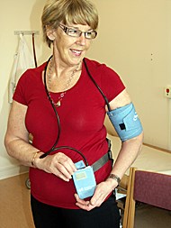Kvinna med kort, ljust hår, glasögon och en röd T-shirt har en blodtrycksmanschett runt vänster arm
