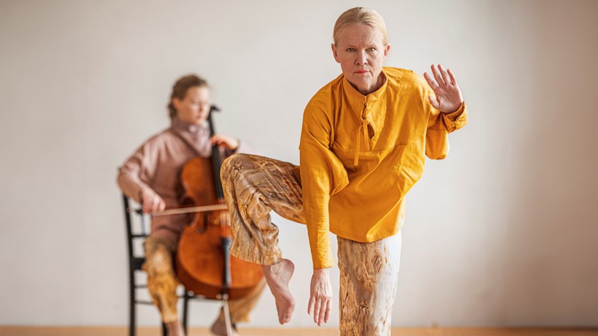 Bilden föreställer två kvinnor. En kvinna står på ett ben i förgrunden. Den andra spelar cello i bakgrunden.