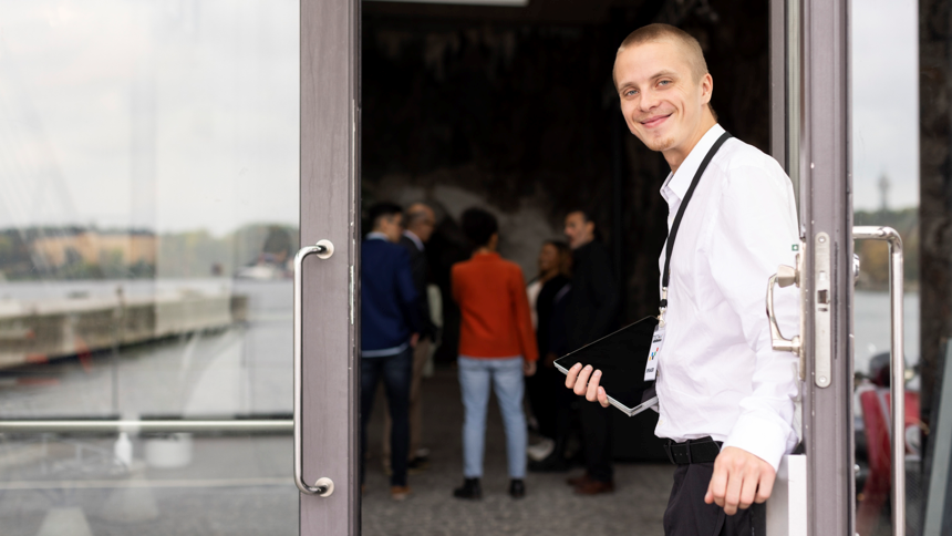 Ung man i vit skjorta och nyckelband runt halsen håller upp ytterdörr